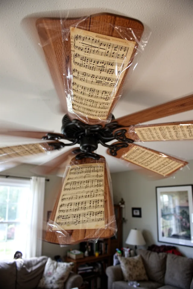 A ceiling fan with wooden blades, each blade wrapped in vintage sheet music pages secured with clear packing tape. The music is visible but slightly yellowed. The fan is on, creating a slight blur. Sunlight from a window, casual living room setting.