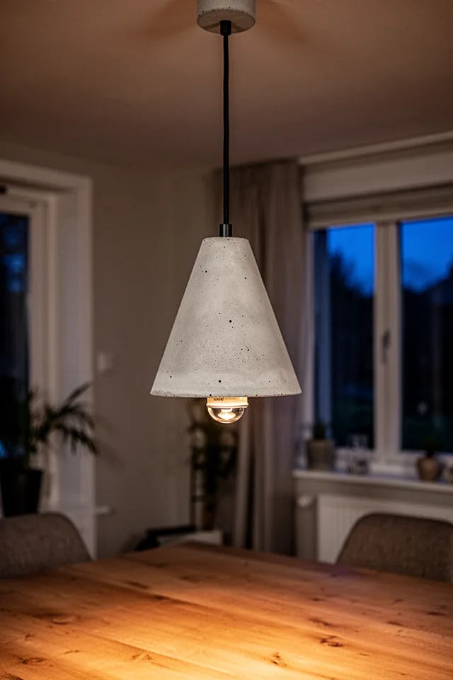 A concrete pendant light hanging from a ceiling over a dining table. The shade is a simple cone shape, 6 inches tall, with a visible socket inside. The concrete is smooth and pale, the cord is black fabric. The light is on, casting a warm glow. Evening setting.