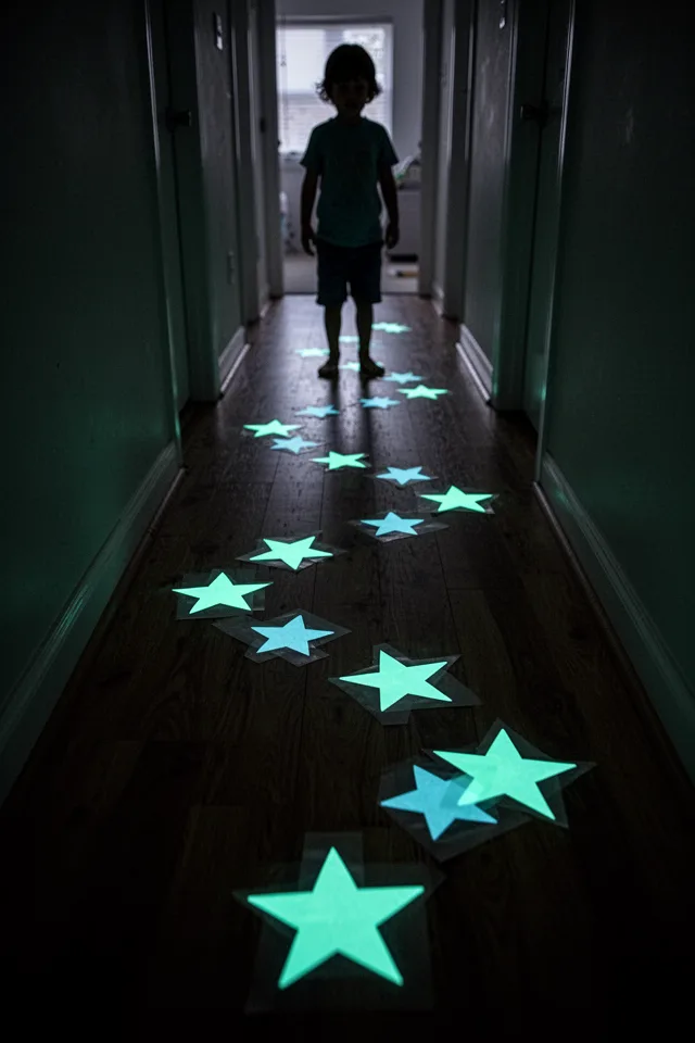 A dark hallway floor with adhesive glow-in-the-dark stars stuck in a winding path. The stars glow a soft greenish-blue. A child