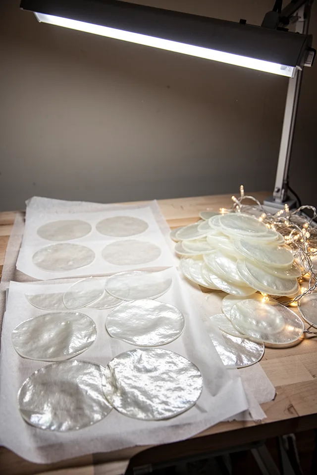 16 Capiz Shell Decor Ideas For A Dreamy Coastal Summer A DIY craft table with parchment paper circles cut out, ironed between wax paper to create a translucent, shell-like texture. Finished "fake capiz" discs are piled next to a strand of lights. Bright overhead craft lighting, neutral background.