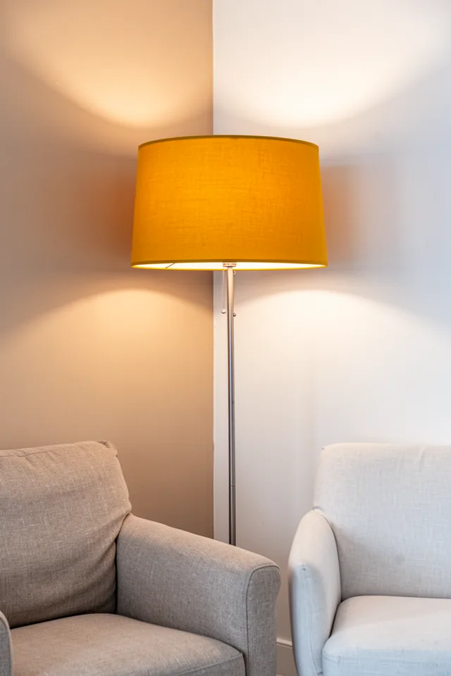 A floor lamp with a large drum shade in bright mustard yellow, placed next to a neutral armchair. The shade casts a warm glow on the wall. The rest of the room is soft beige and white. The lampshade is the only saturated object in that corner.