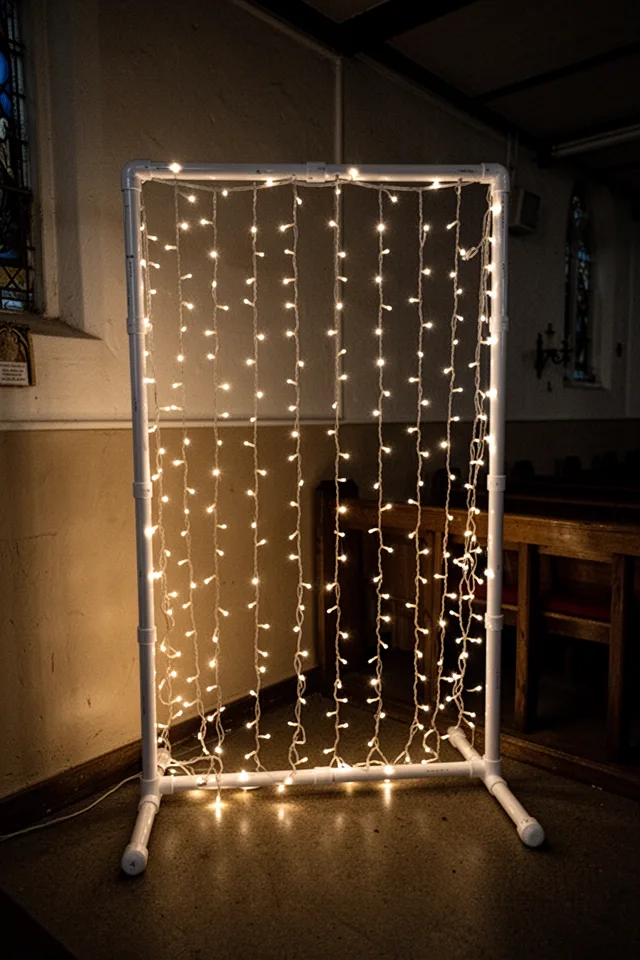 A freestanding frame made of white PVC pipe, draped with a net of warm white LED fairy lights. The lights hang down in vertical strands. The frame stands in a dark corner of a church basement. Soft glow, no other light sources.