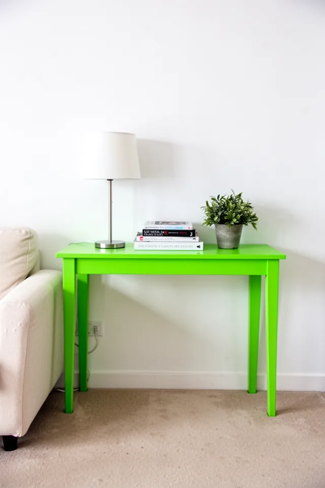 A narrow console table in bright chartreuse green placed against a white wall. On it sits a simple lamp, a stack of books, and a small plant. The green is almost neon but the rest of the room is quiet—cream sofa, beige floor. The console is the only loud element.