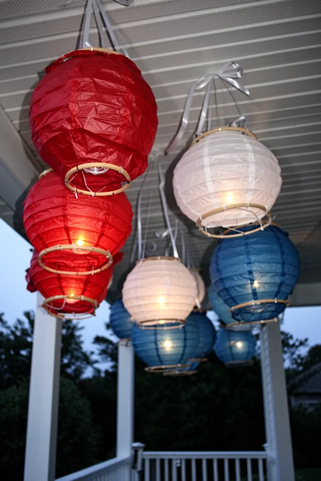 A porch ceiling with several paper lanterns hanging from white ribbon, each lantern in red, white, or blue tissue paper. The lanterns are homemade, slightly imperfect, with wooden dowel frames. Dusk setting, lanterns glowing from within with battery lights.