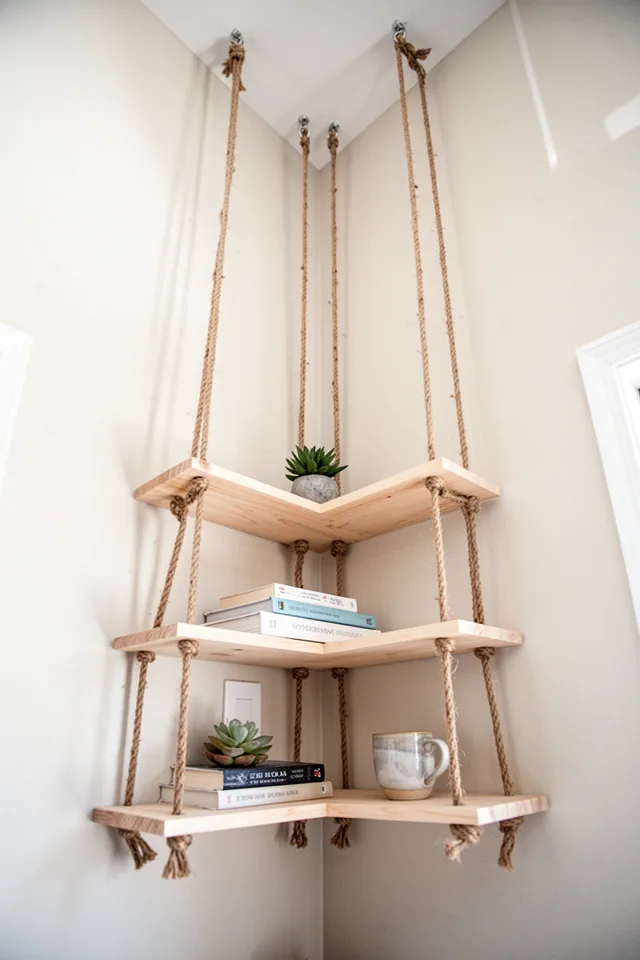 A set of three narrow wooden shelves suspended from the ceiling by ropes in a corner. The ropes are natural jute, the shelves are light birch. The shelves are at staggered heights, and they hold a small succulent, a few paperbacks, and a ceramic cup. The corner has high ceilings, and the rope creates vertical lines. Airy, boho, but restrained. Photo taken from below, looking upward, showing the ropes disappearing into the ceiling.