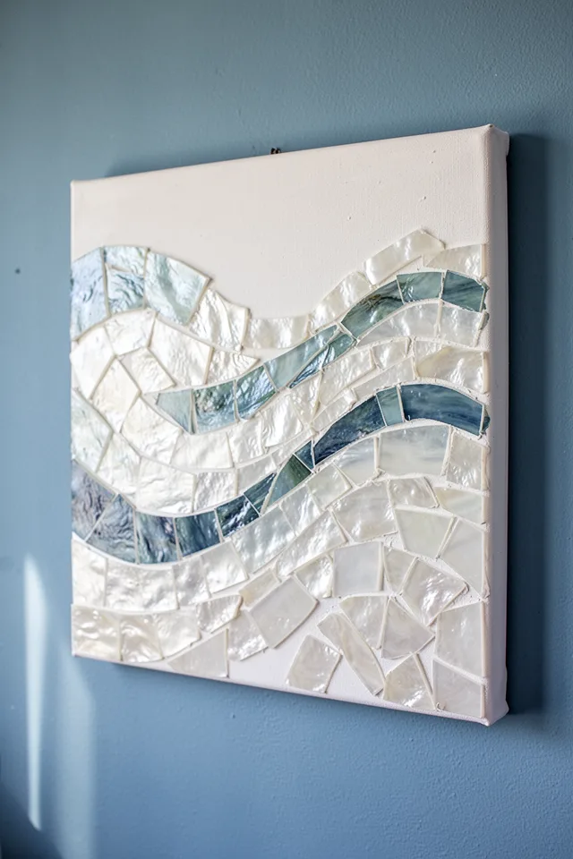 16 Capiz Shell Decor Ideas For A Dreamy Coastal Summer A stretched canvas (16x20 inches) covered with a mosaic of broken capiz shell pieces arranged in an abstract wave pattern. The shells are glued with matte Mod Podge, leaving tiny gaps that show the white canvas below. Hanging on a coastal blue wall. Natural light.