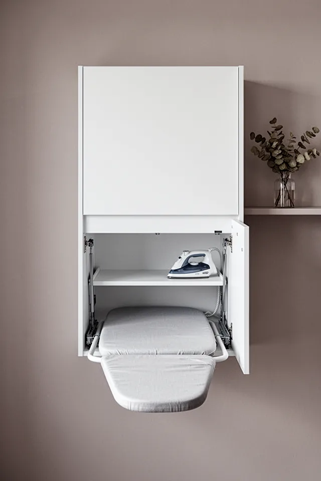 A wall-mounted cabinet with a drop-down ironing board inside. The cabinet door is flush, made of MDF with a white matte finish, and it looks like just another cabinet. Open, the ironing board extends horizontally, revealing a padded surface and a small shelf for the iron. The surrounding wall is a warm light gray. A small vase of dried eucalyptus sits on a shelf next to it. The lighting is soft, indirect. Photographed straight on, slightly elevated, with the cabinet half-open to show the mechanism.