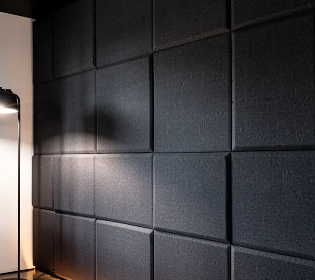 Close-up photograph of a fabric-wrapped acoustic panel wall in a deep charcoal color. The panels are arranged in a stagg