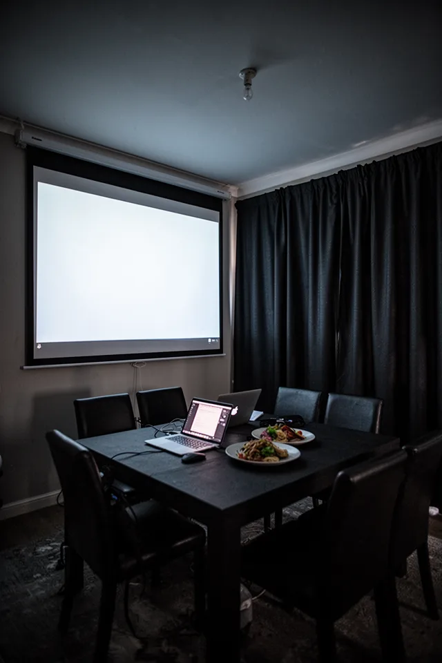Image prompt: A dining room with a projector screen on one wall. Dark blackout curtains drawn fully, deep charcoal with a matte finish. A table in the center with a laptop and plate of food, lit dimly by the screen. The curtains have no sheen, heavy weight. The room is completely dark except for the screen glow. Low angle, wide shot, moody, 24mm lens, high ISO.