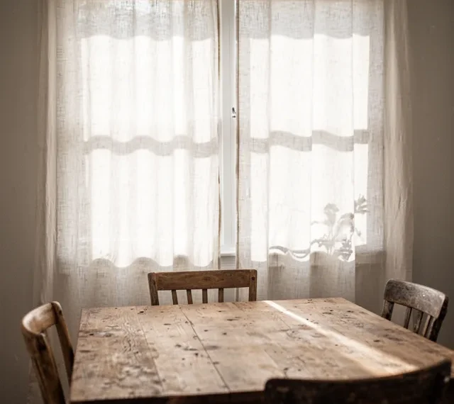Image prompt: A dining room with a weathered wood table and four mismatched chairs. Floor-to-ceiling white linen curtain