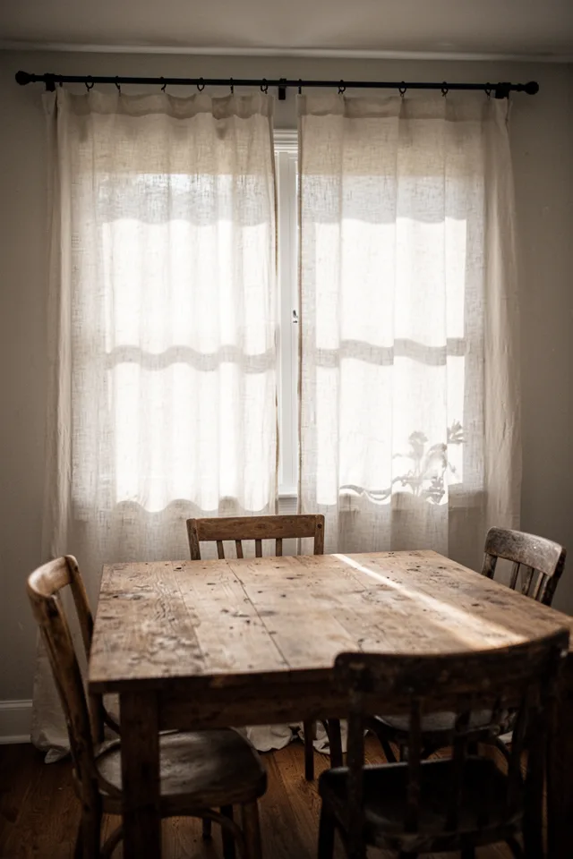 Image prompt: A dining room with a weathered wood table and four mismatched chairs. Floor-to-ceiling white linen curtain panels on a simple black rod. Morning light filters through the linen, casting soft shadows across the tabletop. The fabric has a visible weave and slight unevenness at the hem. No sheers, no hard edges. Shot from a low angle near the table, natural daylight, slightly overexposed to highlight translucency. 35mm lens, warm tone, no people.