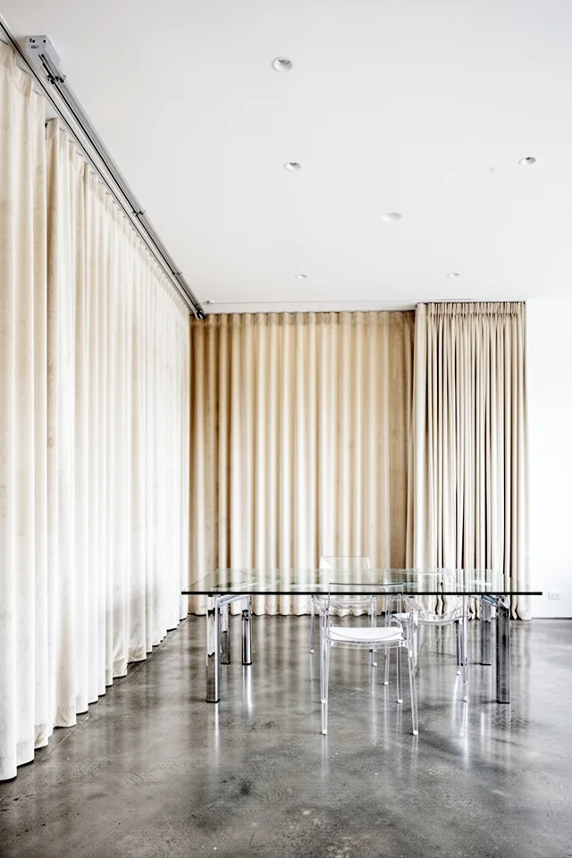 Image prompt: A modern dining room with a white ceiling and minimalist decor. A ceiling-mounted curtain track runs the full length of one wall, with cream velvet panels hanging floor to ceiling. The panels stack neatly at one side. The floor is polished concrete, table is glass with chrome legs. No visible rod. The effect is clean and architectural. Straight-on wide shot, high ceiling, even light.
