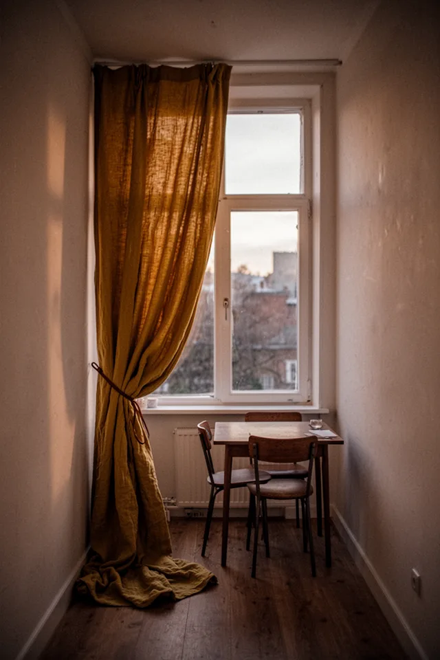 Image prompt: A narrow dining room with a single tall window. A single wide curtain panel is hung on one side only, gathered loosely and tied back with a leather cord. The panel is a rich mustard yellow cotton-linen, pooling slightly on the floor. The other side of the window is bare. A small table with two chairs is placed asymmetrically under the window. Warm evening light. 35mm lens, editorial, slightly moody.