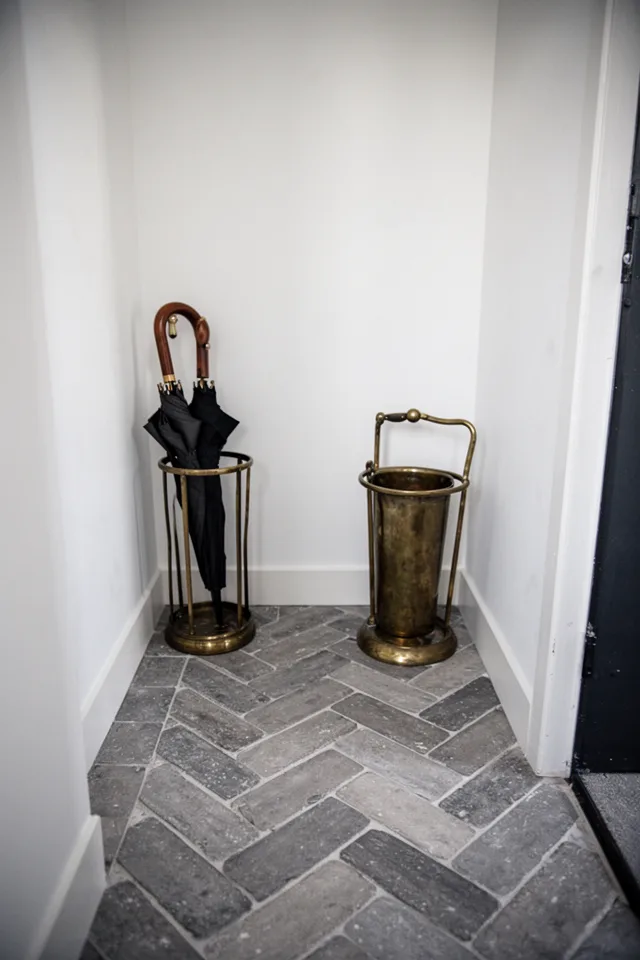 Image prompt: Small entryway, gray brick-look tile (3x9 inch) laid in a herringbone pattern. The tile has a slight tumbled edge, soft matte finish. A brass umbrella stand, stark white walls. Lens: 35mm, mood: French industrial, grounded. No people.